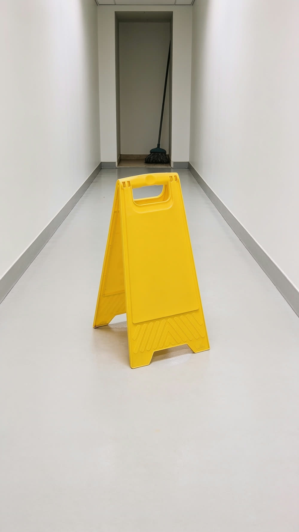 A bright yellow folding wet floor sign standing dead centre of an empty office corridor, shot from a low hero angle, the floor around it completely dry.