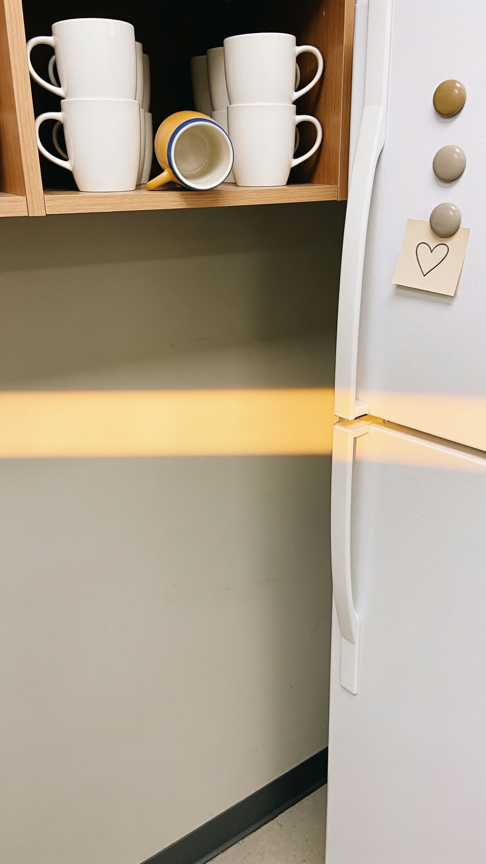 A quiet stretch of breakroom wall with a warm band of golden light crossing it, an open shelf of white mugs with one yellow mug turned upside down among them, and a small hand-drawn heart on a note pinned to the fridge.