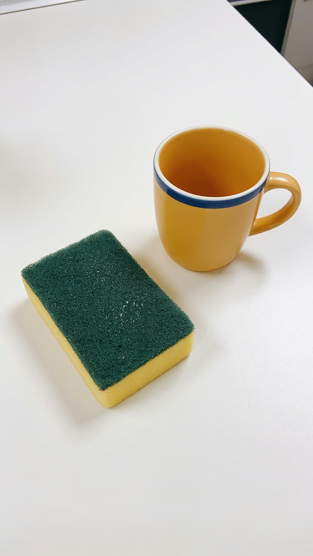 A straight overhead flat-lay of a kitchen counter: a visibly damp yellow and green sponge posed dead square beside a warm yellow coffee mug with a thin blue band, arranged like museum pieces on an otherwise bare counter.