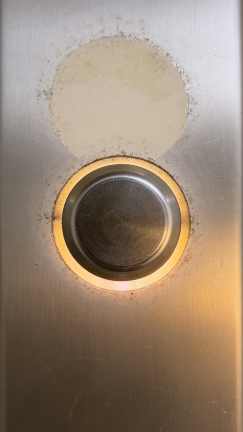 An extreme close-up of the elevator's call button plate: a round illuminated up button, its edges dulled and smudged from years of pressing, mounted above a plain unlit down button on brushed steel.